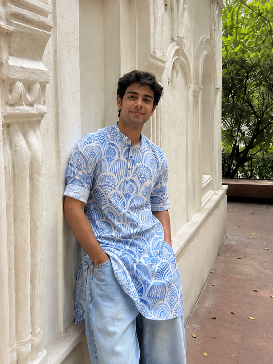Man wearing a blue patterned shirt leaning against a white architectural wall.