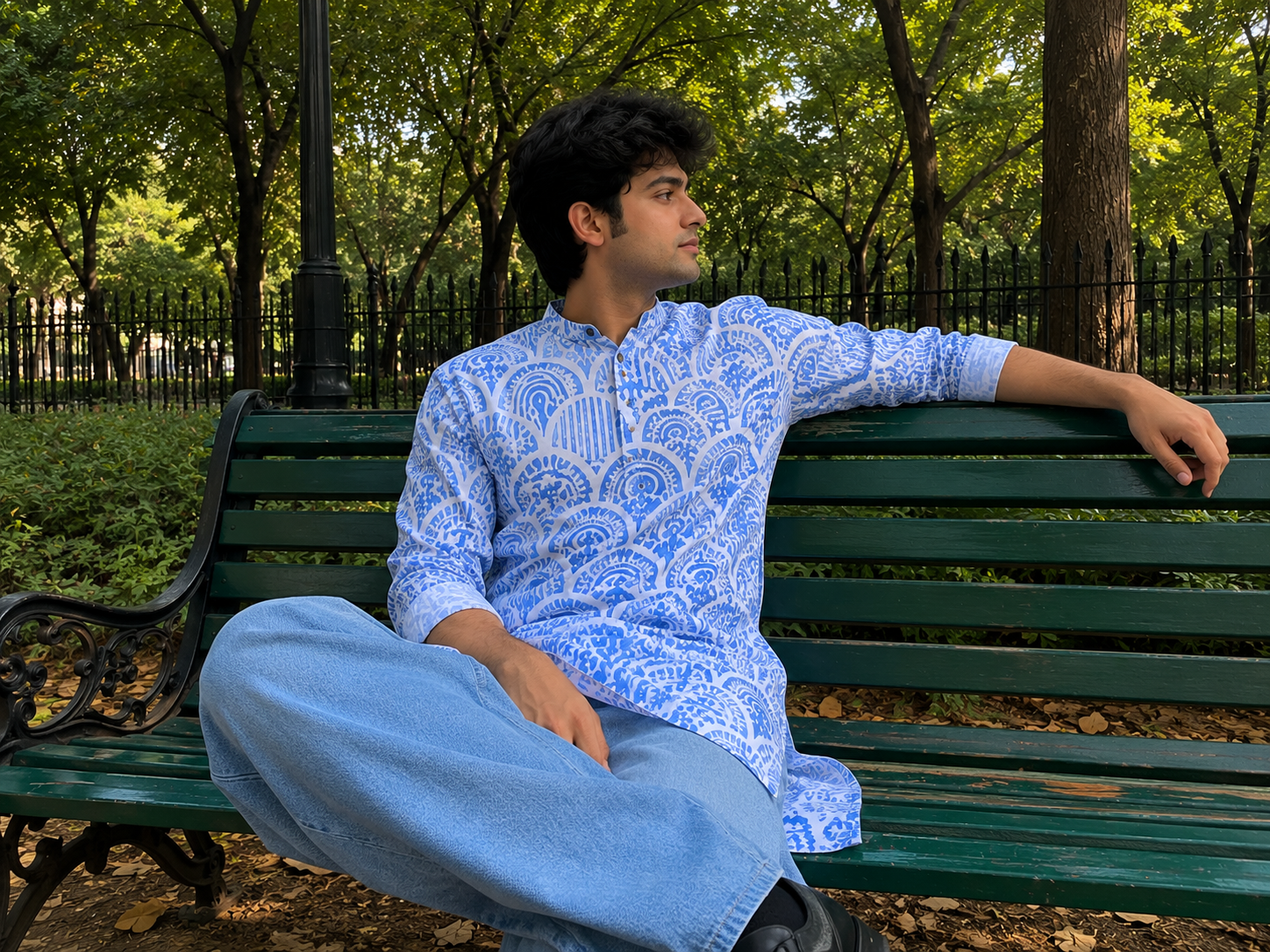 Man sitting on a park bench wearing a blue patterned shirt and light blue jeans.
