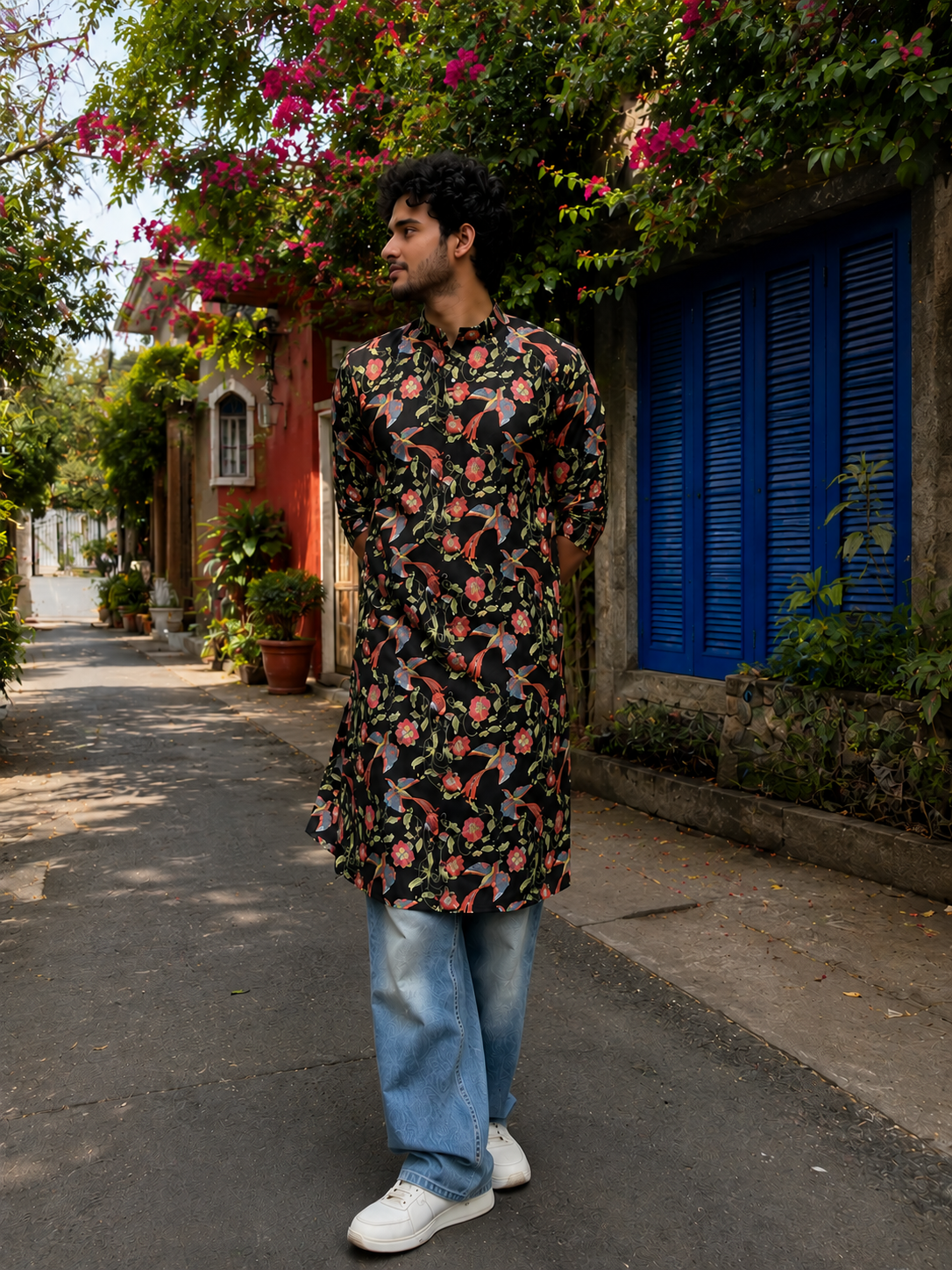 Man wearing a floral kurta with jeans and white sneakers standing on a street with plants and flowers.
