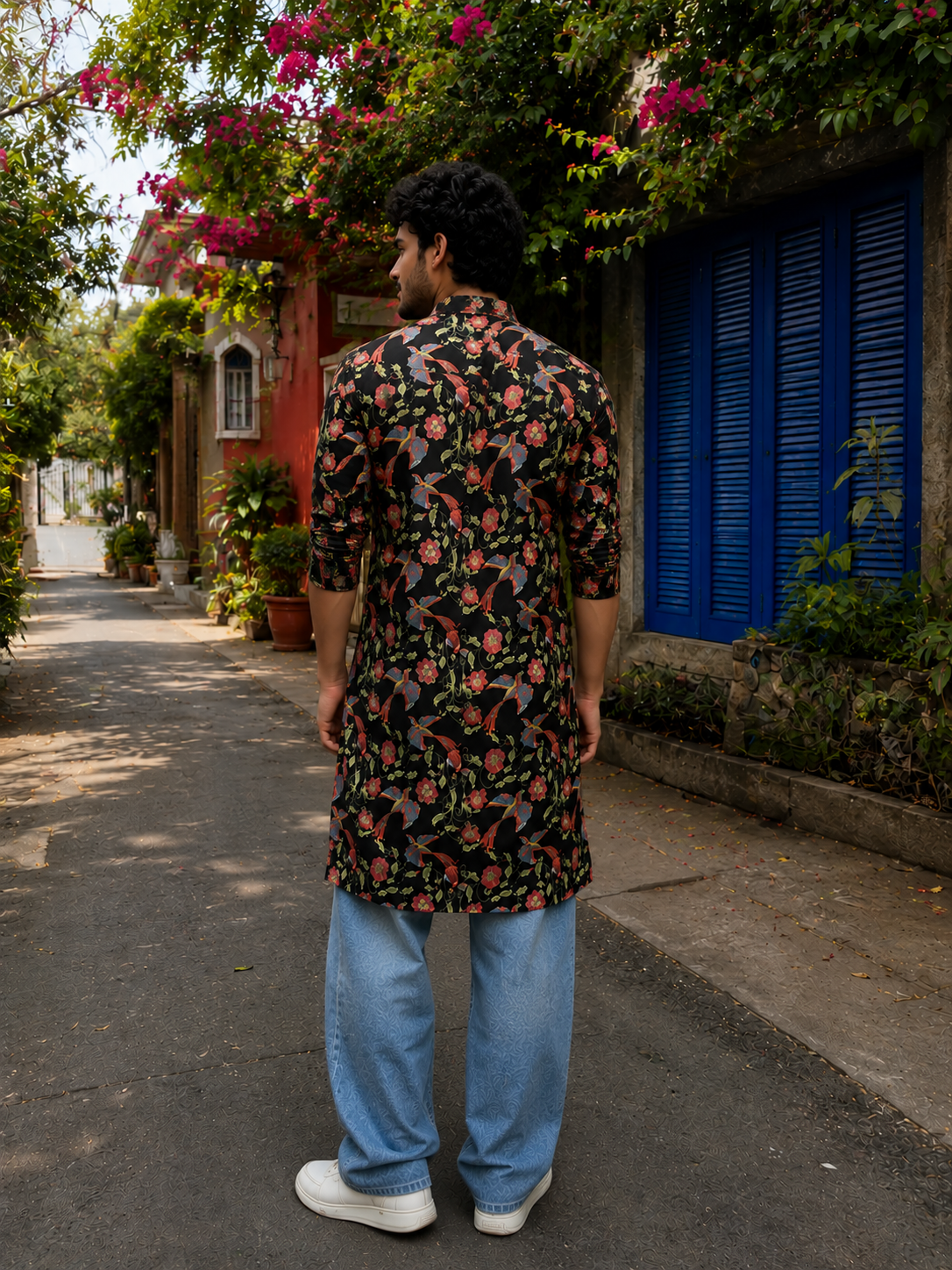 Man wearing a floral shirt and blue jeans walking down a street with colorful buildings and plants.