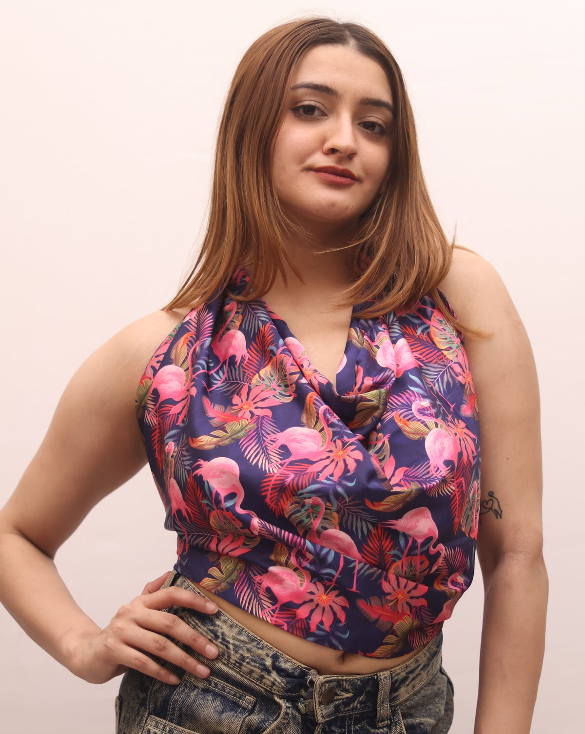 A woman posing in a sleeveless halter crop top with a vibrant pink, purple, and blue flamingo print, paired with high-waisted, faded blue jeans against a plain light background.