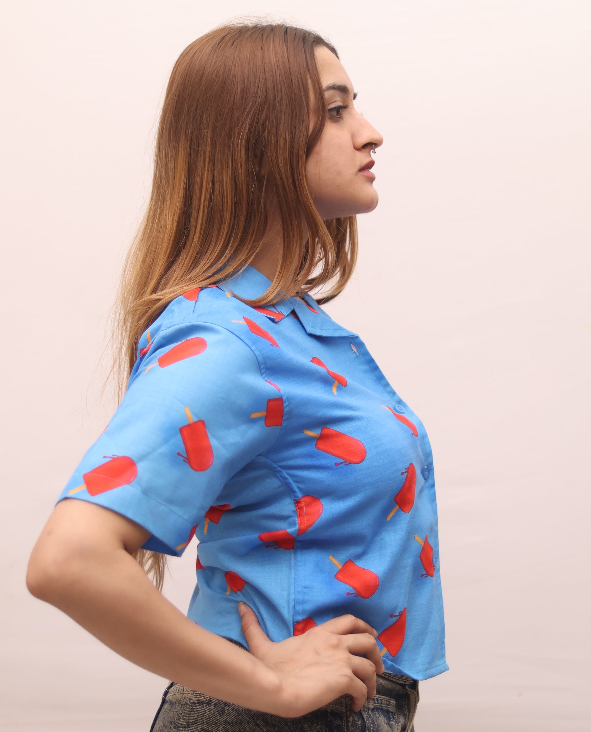 A woman in profile wearing a light blue short-sleeve button-up shirt with a red popsicle print, paired with gray denim jeans, against a plain light beige background.