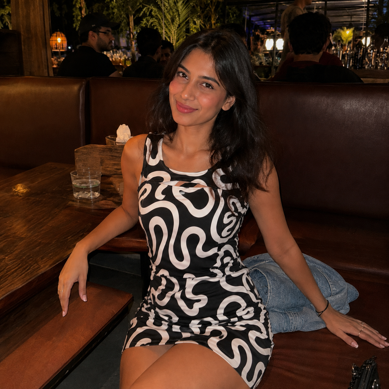 A woman sitting in a restaurant booth wearing a sleeveless black mini dress with a bold white abstract wavy pattern. She is smiling and looking at the camera, with a denim jacket resting on the booth next to her.