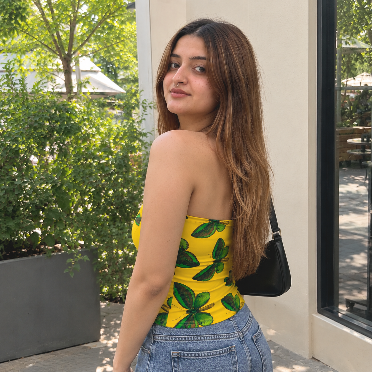 A woman wearing a bright yellow strapless tube top with a green leaf pattern, paired with light blue jeans and a black shoulder bag, standing outdoors next to a building.