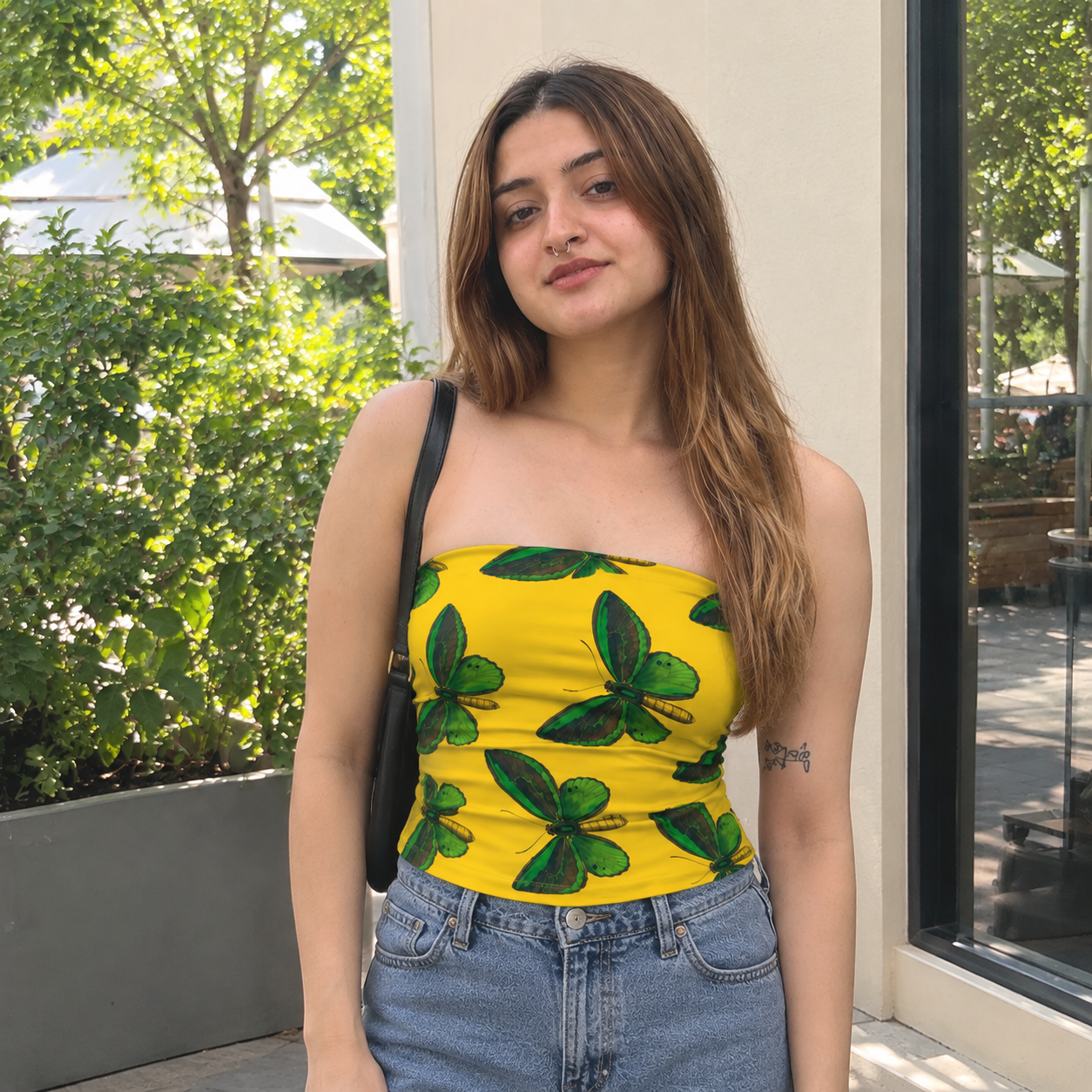 A woman wearing a bright yellow strapless tube top with a green butterfly print, paired with light-wash blue jeans and a black shoulder bag, standing outdoors in front of a building with greenery.