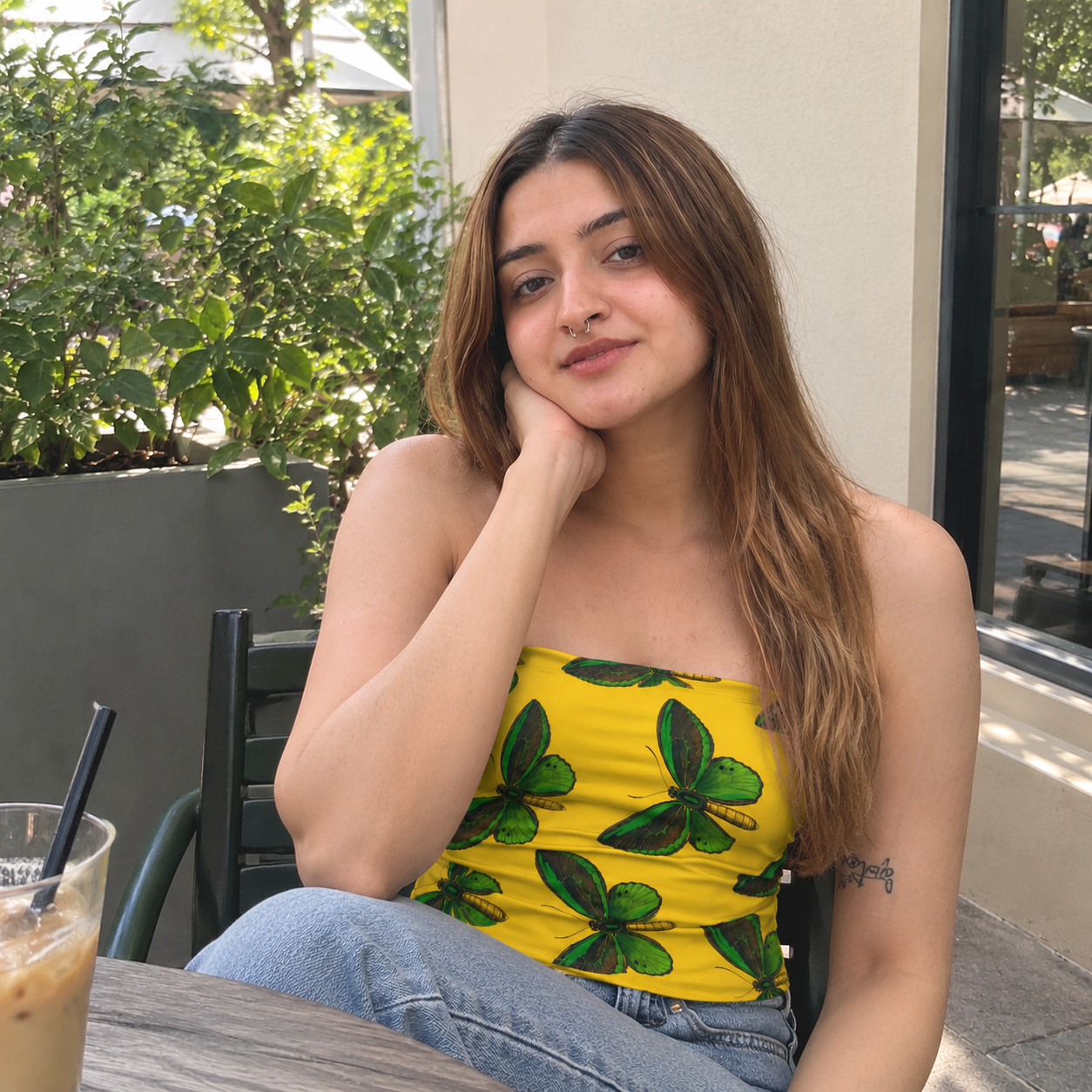 A woman sitting at an outdoor cafe table wearing a bright yellow strapless tube top with a green butterfly pattern, paired with light blue jeans.