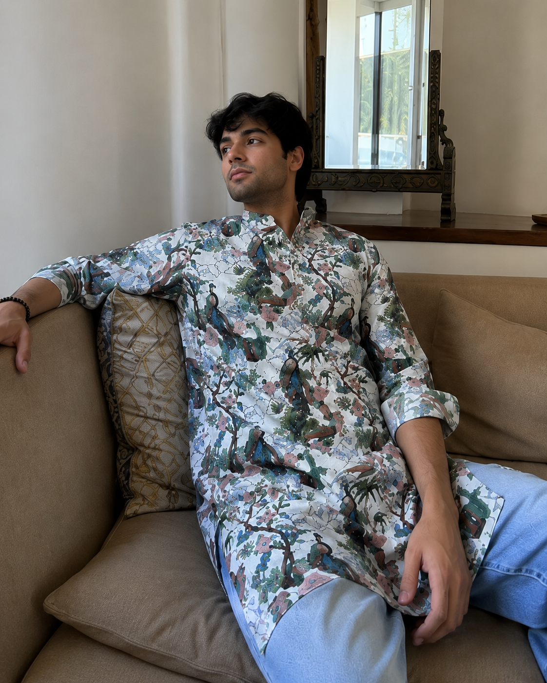 Man wearing a floral shirt sitting on a couch in a room with a mirror on the wall.
