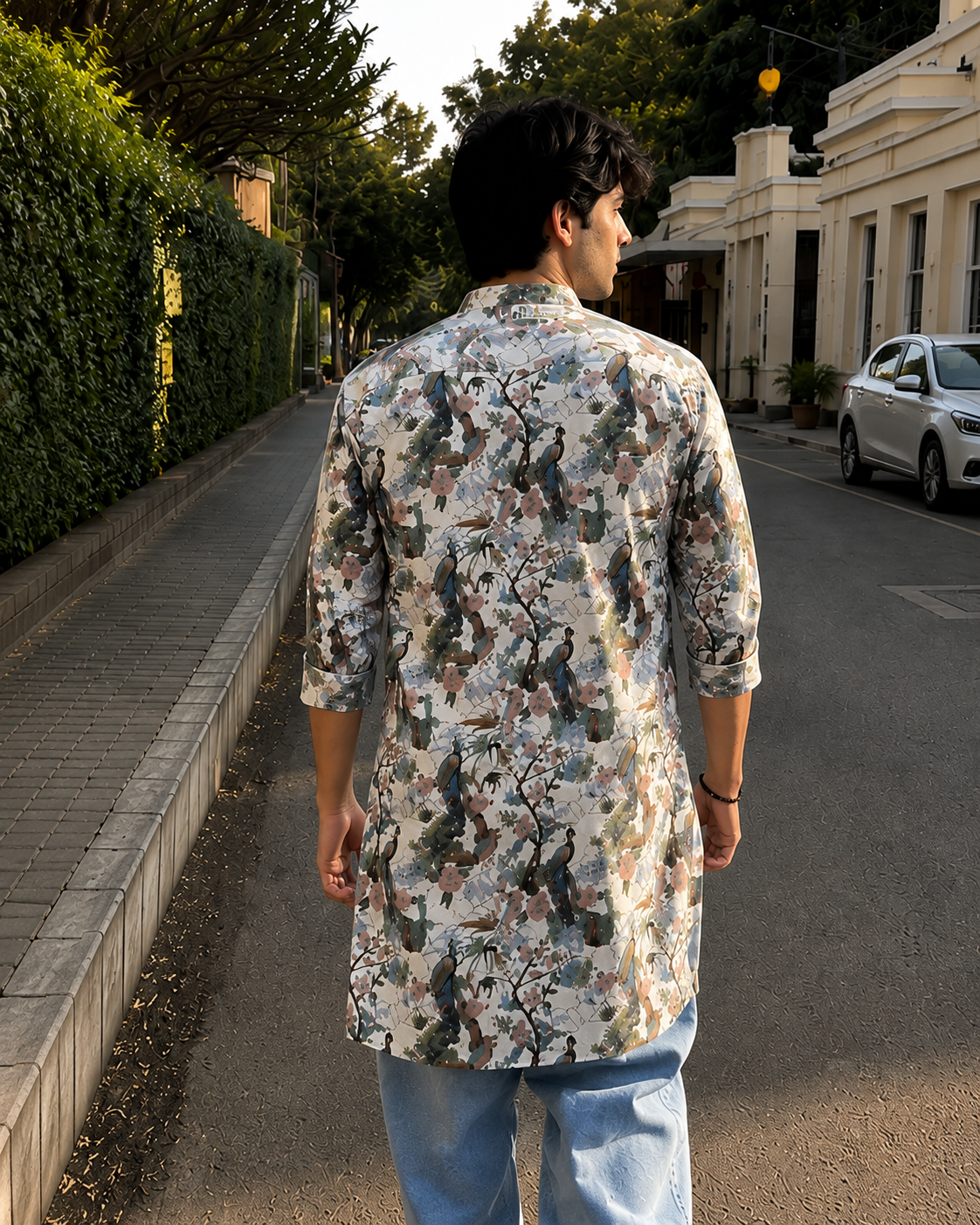 Man walking on a sidewalk wearing a patterned kurta and jeans, with a building and car in the background.