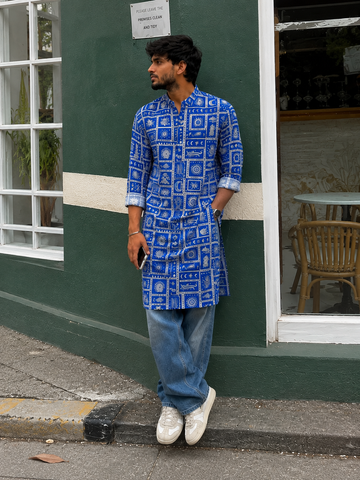 Man wearing a blue patterned kurta with jeans and white shoes standing against a green wall.