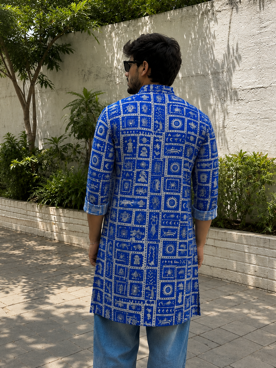 Man wearing a blue patterned shirt standing outdoors with a white wall and greenery in the background