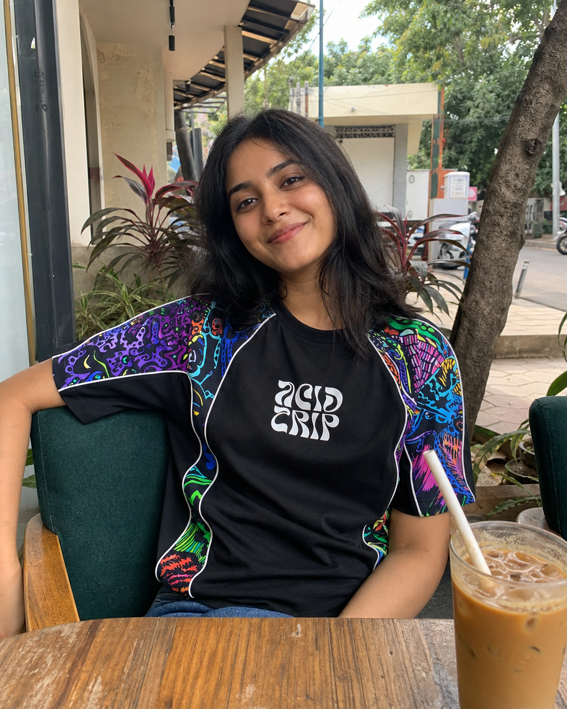 Person wearing a colorful jacket and black shirt with text, sitting outdoors with iced coffee.