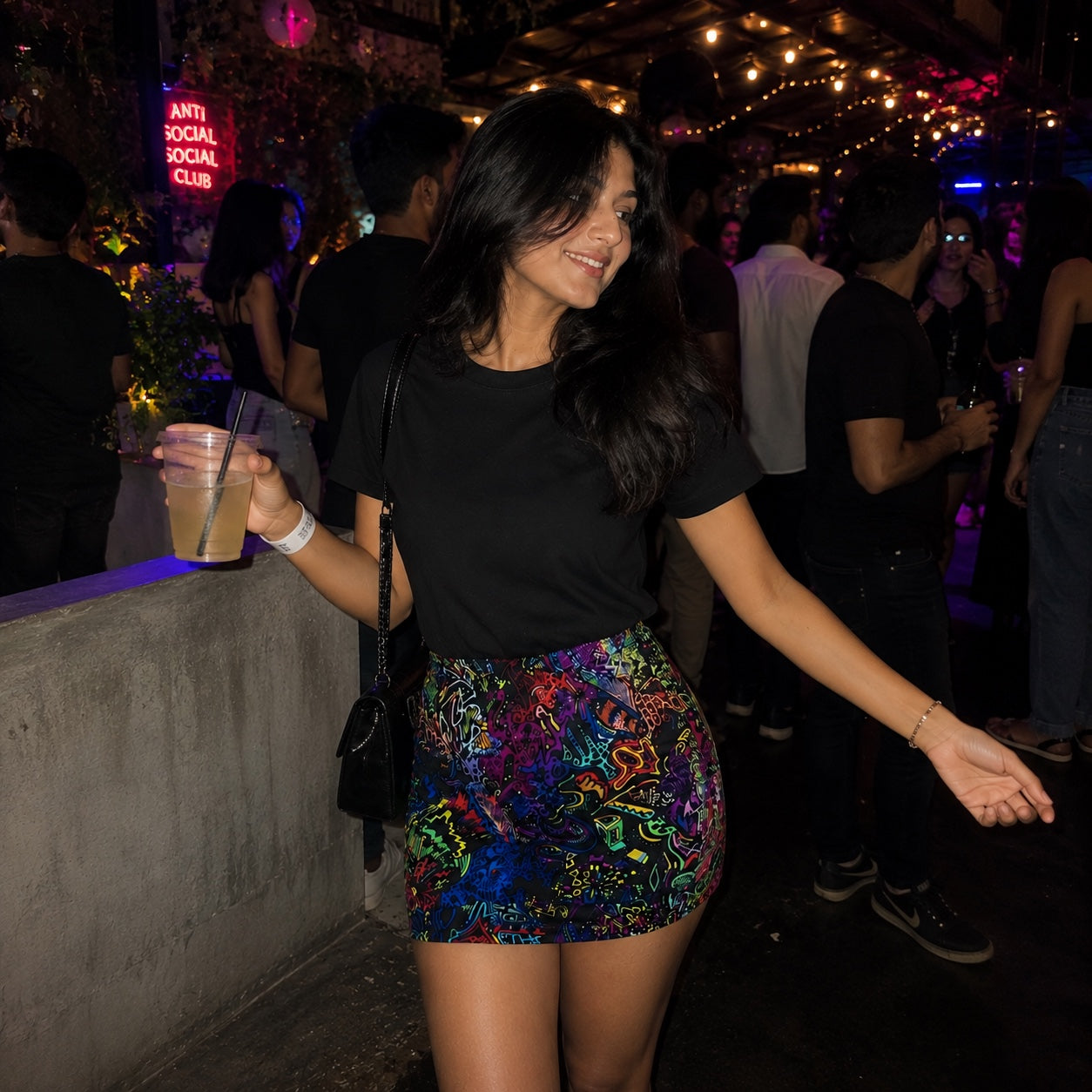 Woman in a colorful acid trip and black top at a lively event with neon signs and people in the background.