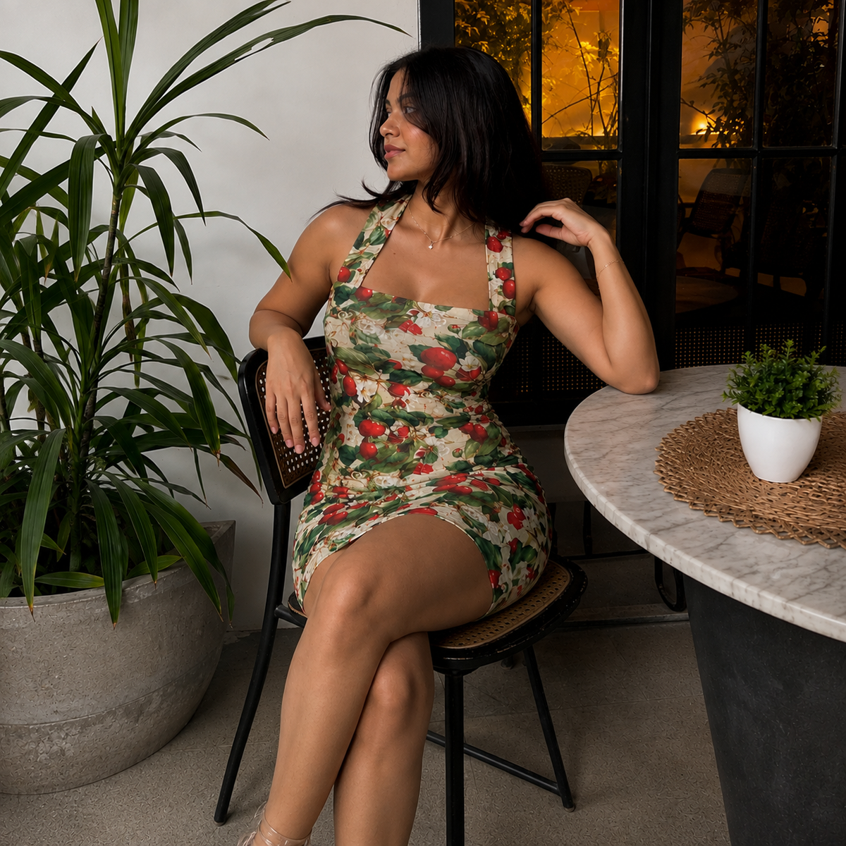 A woman sits on a black chair in a modern indoor setting, wearing a fitted halter mini dress with a red and green floral print on a beige background. A large potted plant is on the left, and a marble-topped table with a small plant is on the right.