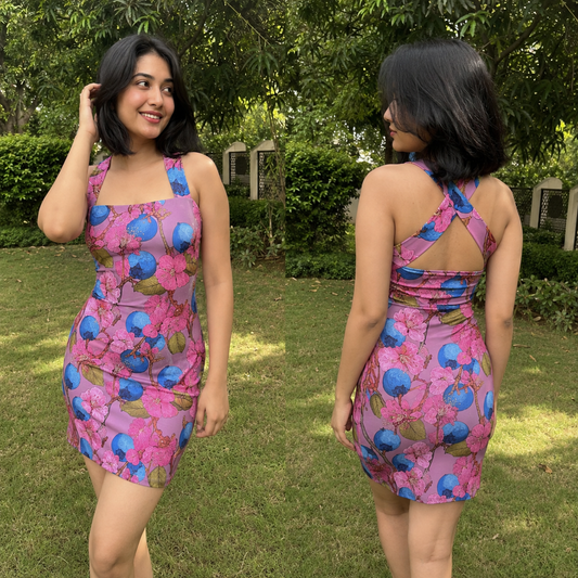 A woman modeling a fitted mini dress in an outdoor garden setting, shown from both the front and back. The dress has a vibrant pink and purple floral pattern with blue berry-like motifs and green leaves.