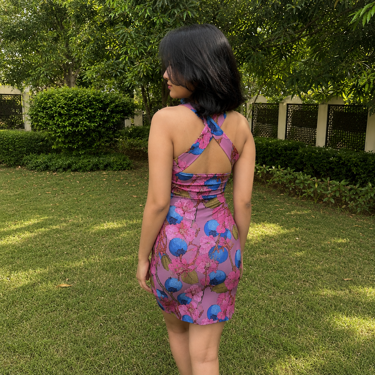 A woman standing outdoors on a grassy lawn, viewed from behind, wearing a sleeveless pink mini dress with a blue and green floral print and an open back.