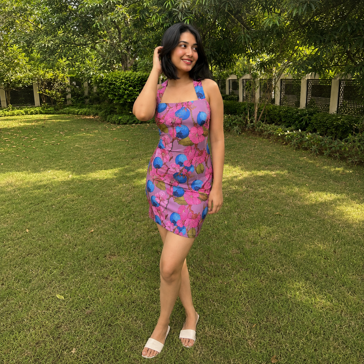 A woman wearing a pink and blue floral print mini dress with a corset-style bodice, standing on a green lawn in a garden setting.