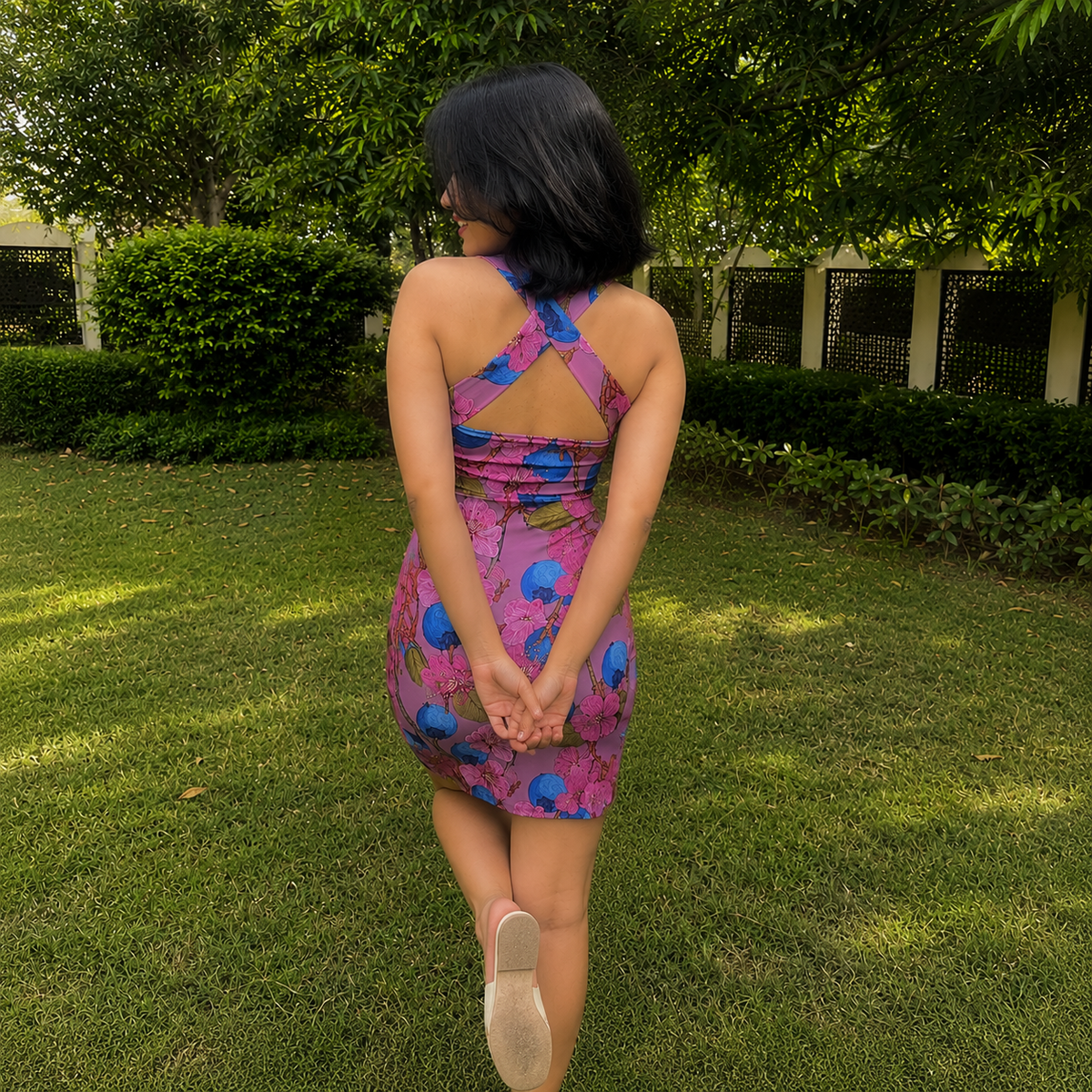 A woman is photographed from behind outdoors on a grassy lawn, wearing a fitted mini dress with a pink and purple base and blue abstract floral-like print. The dress has a backless open back with crisscross straps.