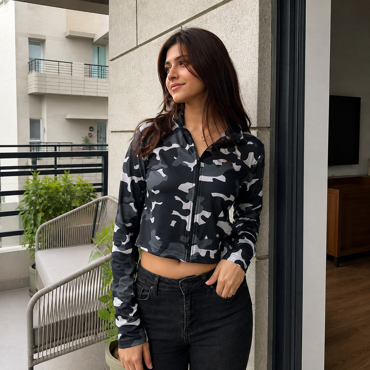 A woman wearing a black and white camouflage patterned zip-up crop jacket and black jeans, standing on a balcony.