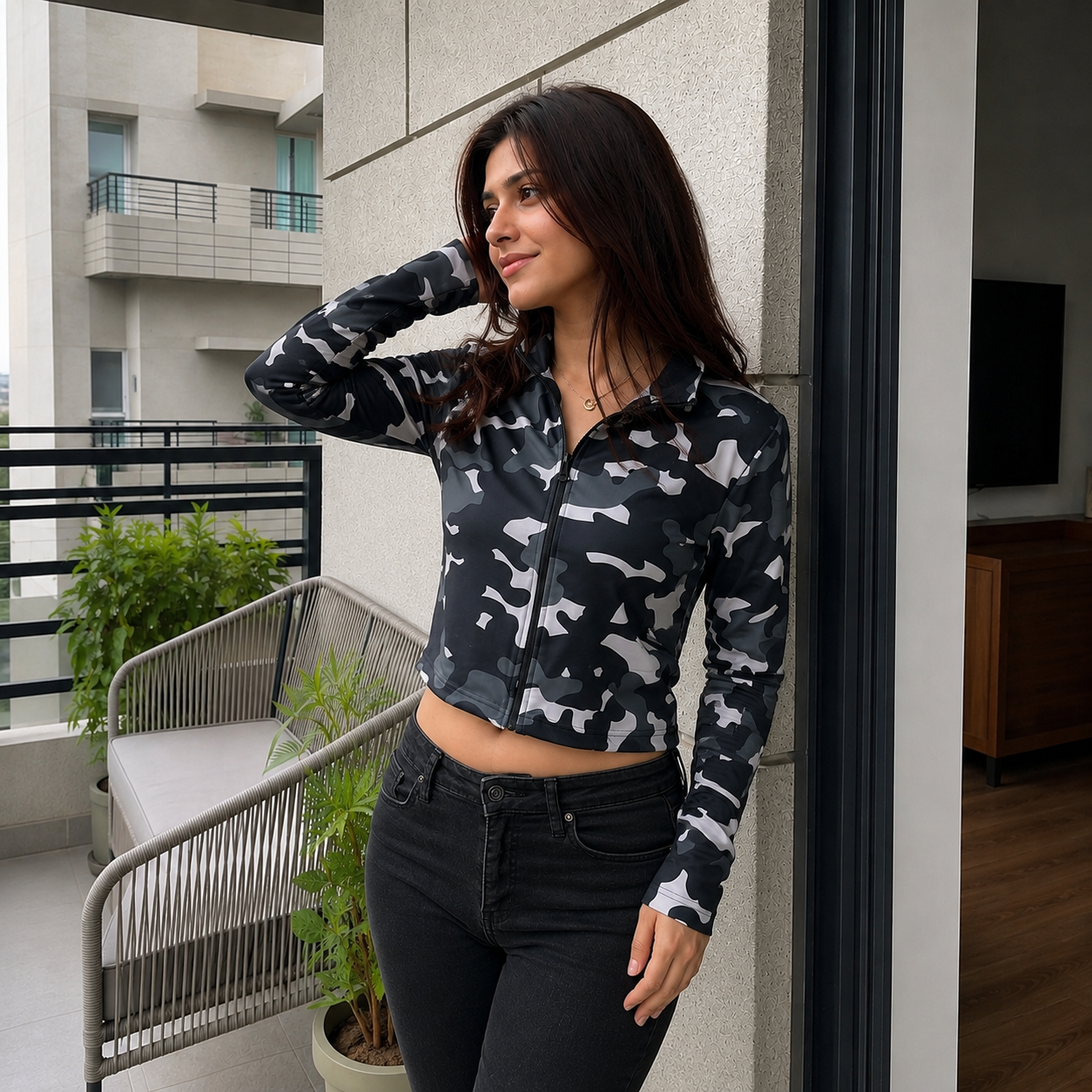A woman wearing a black and gray camouflage patterned long-sleeve zip-up crop top, standing on a balcony next to a modern glass door.