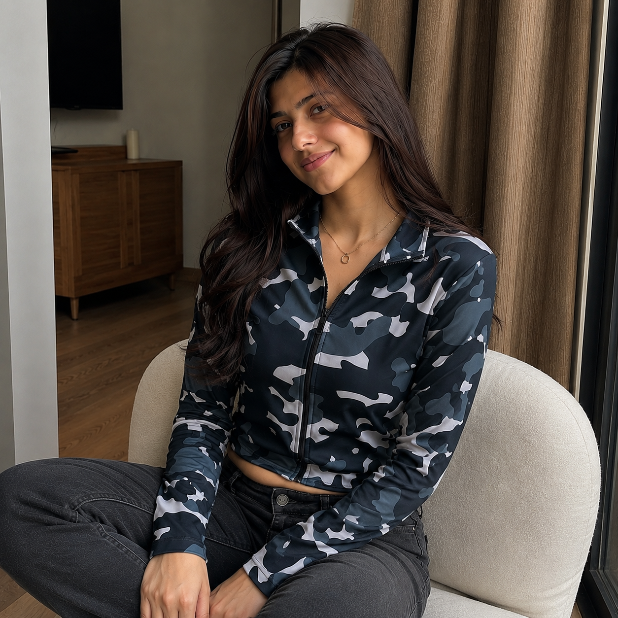 A woman sitting on a beige armchair wearing a dark camouflage zip-up jacket and dark gray jeans in a living room setting.