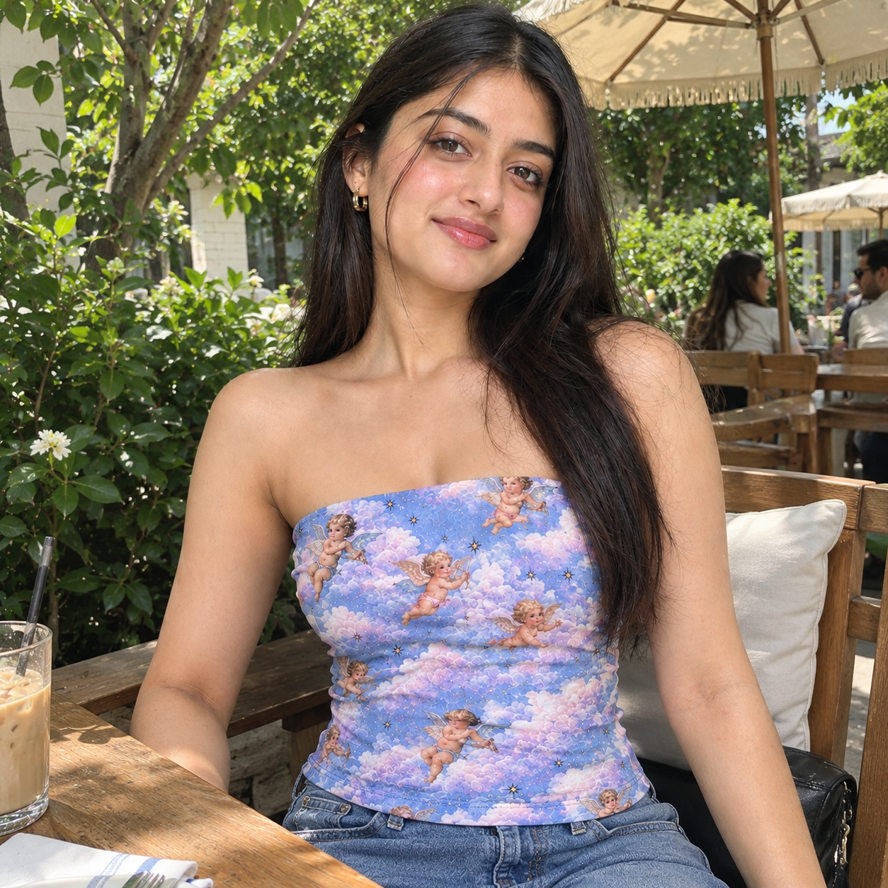 A woman sitting at an outdoor cafe table wearing a strapless tube top with a pastel blue and purple cherub and cloud print, paired with blue jeans.
