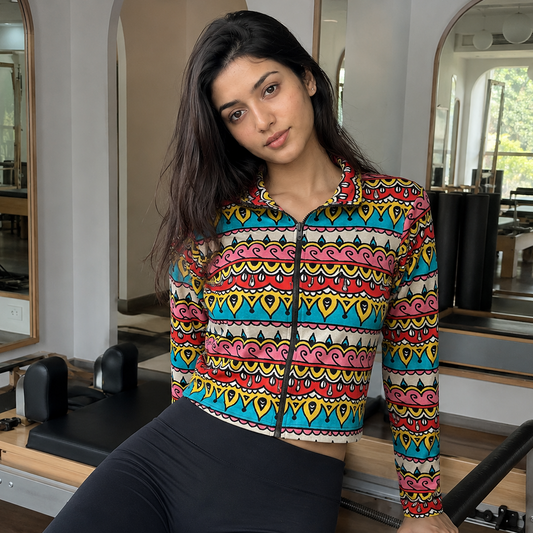 A woman posing in a fitness studio wearing a colorful, long-sleeve cropped zip-up jacket with a vibrant geometric pattern and black leggings.