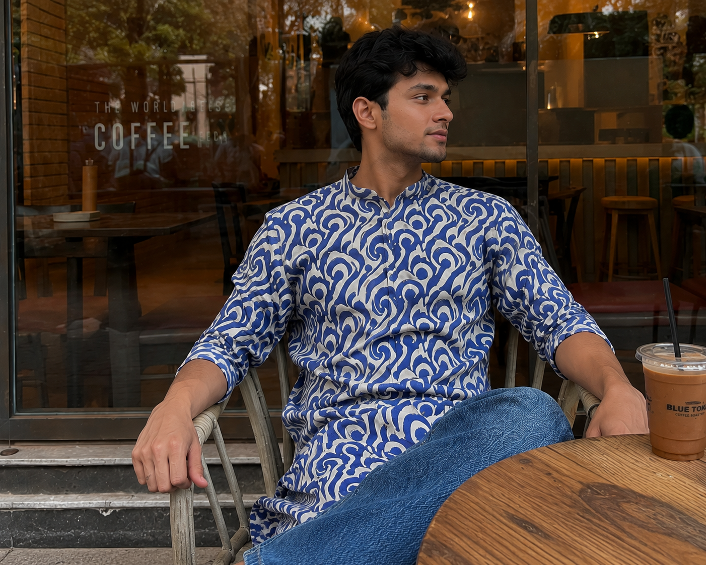 Man sitting at a table in a cafe wearing a blue patterned shirt.