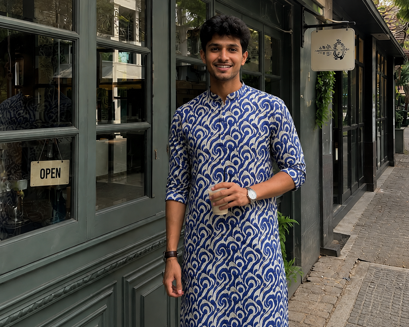 Man in a blue and white patterned kurta standing outside a building with an 'Open' sign.