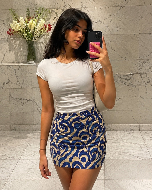 Woman taking a mirror selfie wearing a white top and patterned skirt in a bathroom.