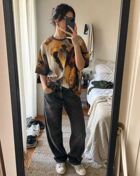 Person taking a mirror selfie wearing a graphic t-shirt and dark jeans in a bedroom.