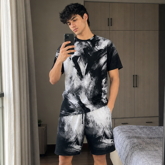 Man taking a mirror selfie wearing a black and white tie-dye outfit in a bedroom.