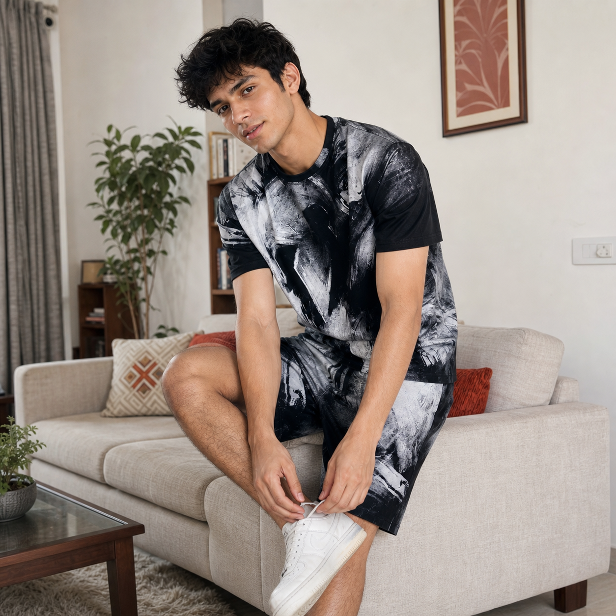 Man sitting on a couch wearing a black and white patterned t-shirt and white sneakers in a living room.