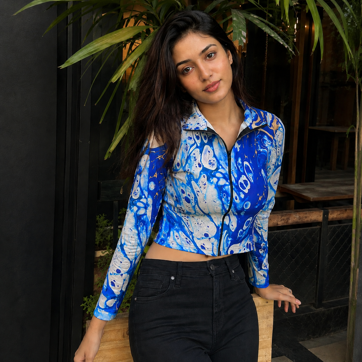 A woman wearing a blue and white abstract patterned long-sleeve crop top with a mock zipper, paired with black jeans, posing in front of a dark doorway with green plants.