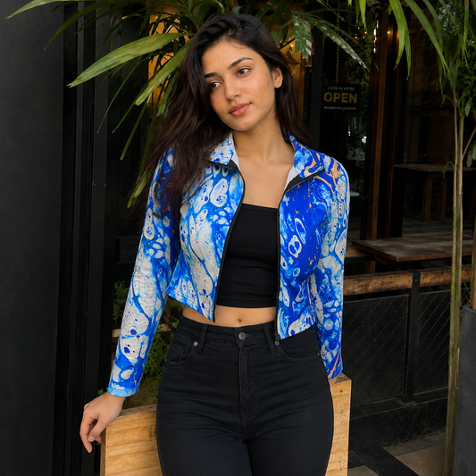 A woman wearing a blue and white abstract patterned zip-up jacket over a black crop top and black jeans, standing in front of a storefront with plants.