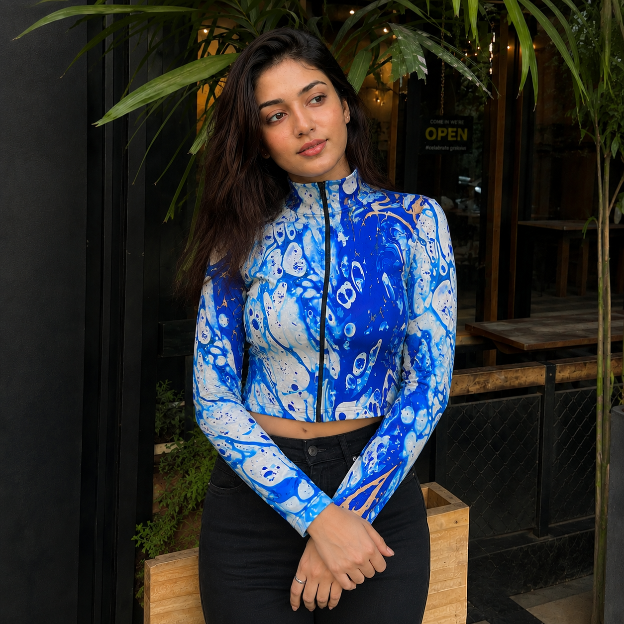 A woman wearing a long-sleeve mock-neck crop top with a blue, white, and black abstract pattern, paired with black pants, standing in front of a storefront with green plants.