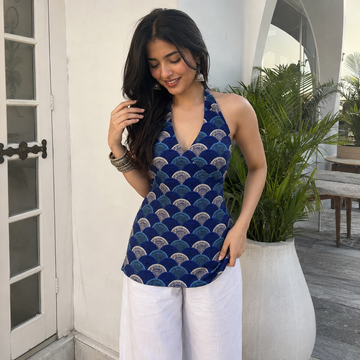 A woman wearing a blue halter top with a repeating scallop pattern in navy, teal, and beige, paired with white pants, standing in front of a white wall with a large potted plant.