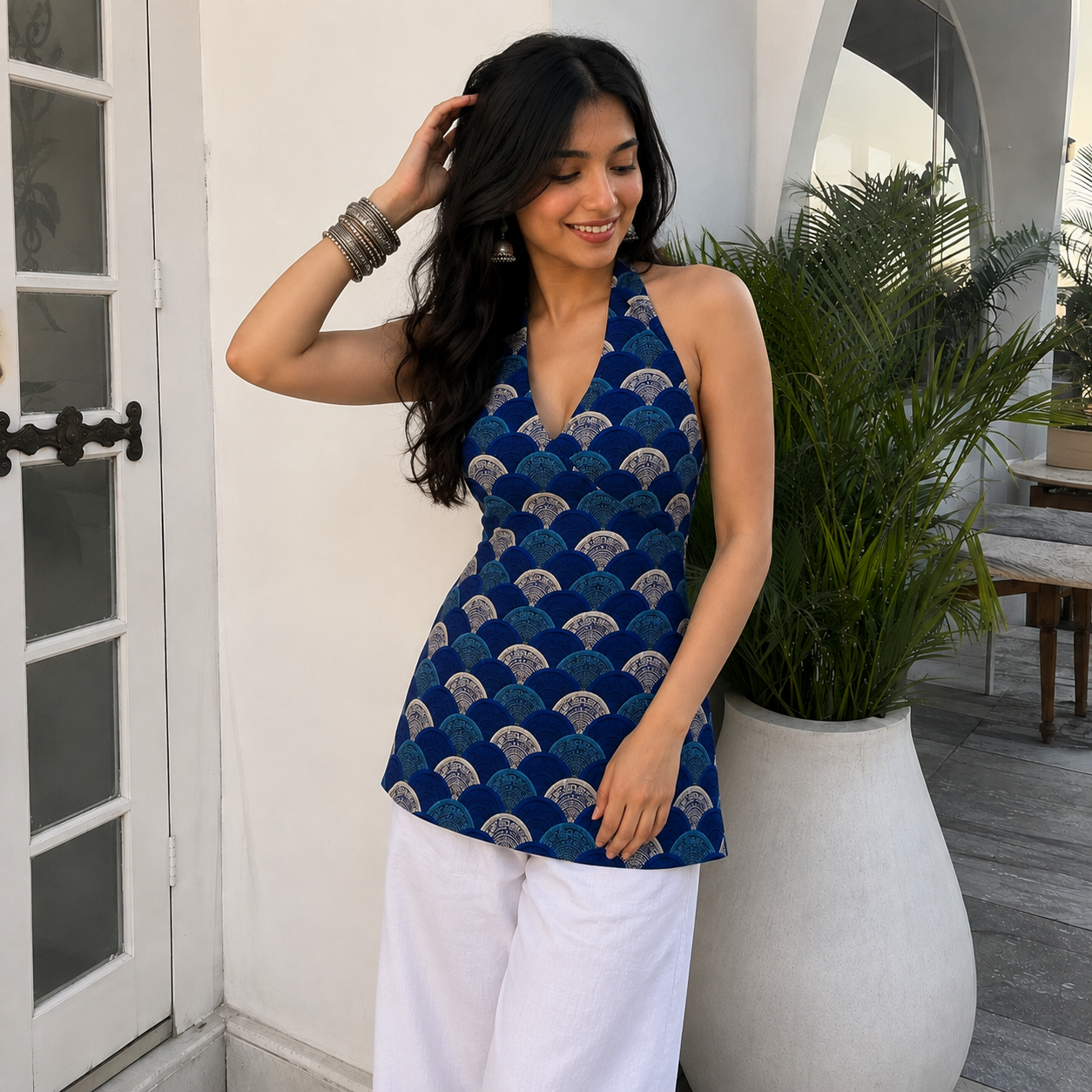 A woman wearing a blue halter top with a repeating beige and blue fan pattern, paired with white pants, standing in a bright, modern outdoor patio setting.