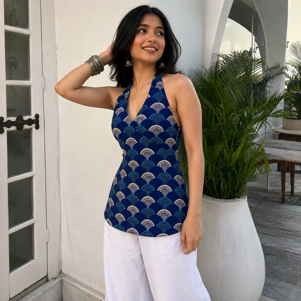 A woman wearing a navy blue halter top with a repeating gold and white fan pattern, paired with white pants, posing outdoors next to a large potted plant.