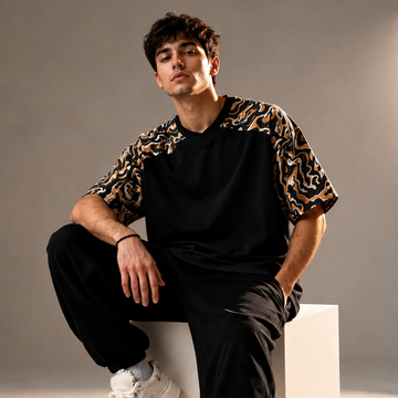 Man sitting on a white cube wearing a black and leopard print shirt with black pants and white sneakers.