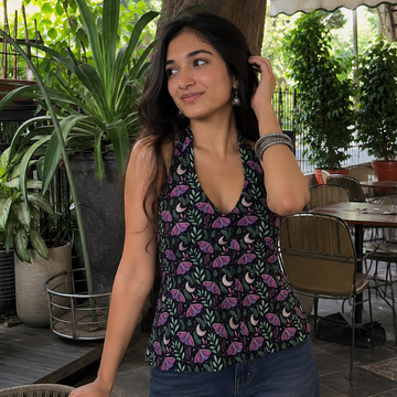 A woman wearing a sleeveless dark green halter top with a purple and black floral print, paired with blue jeans, standing in an outdoor patio setting with plants and tables.