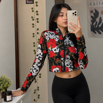A woman takes a mirror selfie wearing a black long-sleeve crop top with a red and white floral and skull pattern, paired with black leggings, in front of a large wooden-framed mirror.