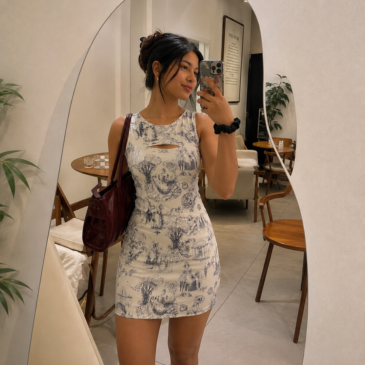 A woman takes a mirror selfie in an indoor boutique setting, wearing a sleeveless bodycon mini dress with a blue toile-style print on a cream background and a small chest cutout. She is carrying a dark burgundy shoulder bag.