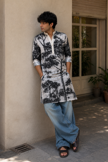 Person wearing a black and white floral kurta with blue pants standing against a beige wall.